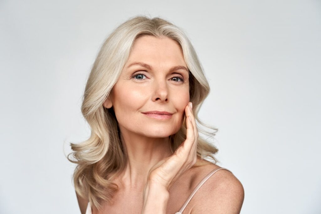 A mature woman with fair skin and blonde hair smiles softly, touching her cheek gently. She exudes a sense of confidence and well-being, highlighting the potential benefits of cosmetic dermatology in achieving youthful, glowing skin.